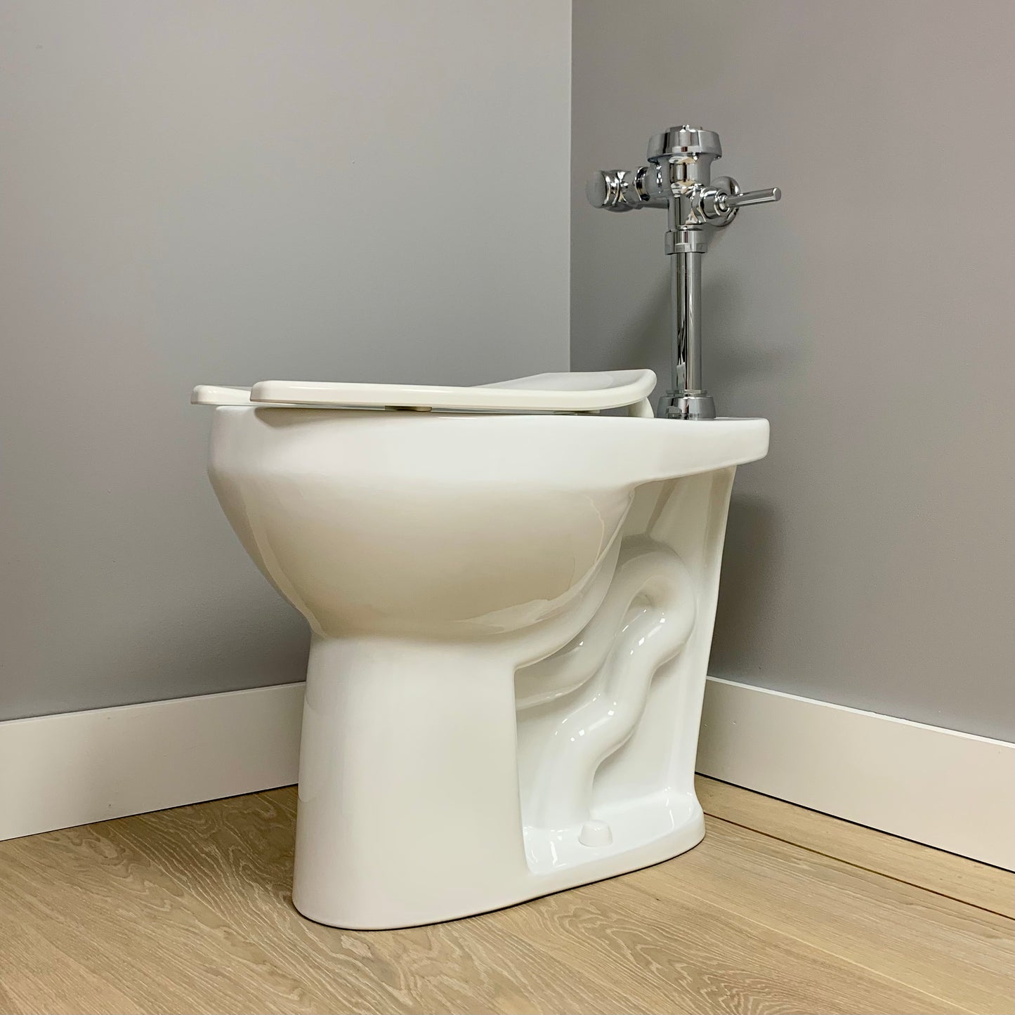 A white, extra tall elongated commercial toilet bowl with an open lid, mounted on the floor against a gray wall.