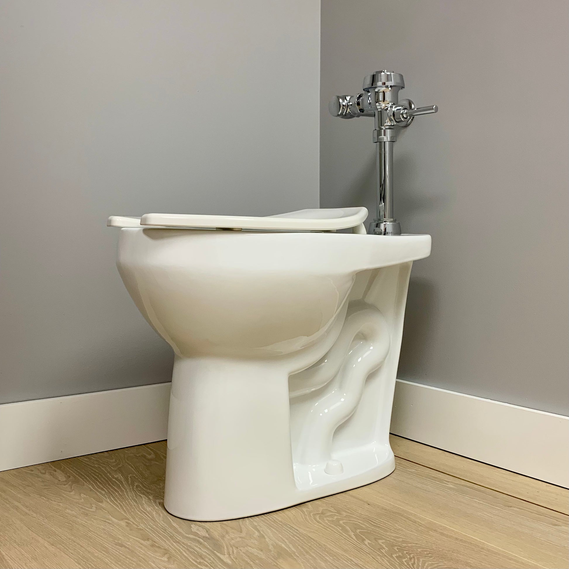A white, extra tall elongated commercial toilet bowl with an open lid, mounted on the floor against a gray wall.