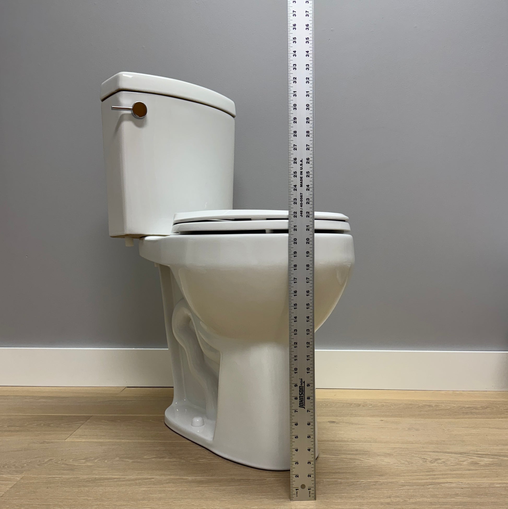 White toilet with a measuring tape against a gray wall and wooden floor